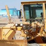 CAT 160K Motor Grader front work lights and blade lift cylinders at an industrial site